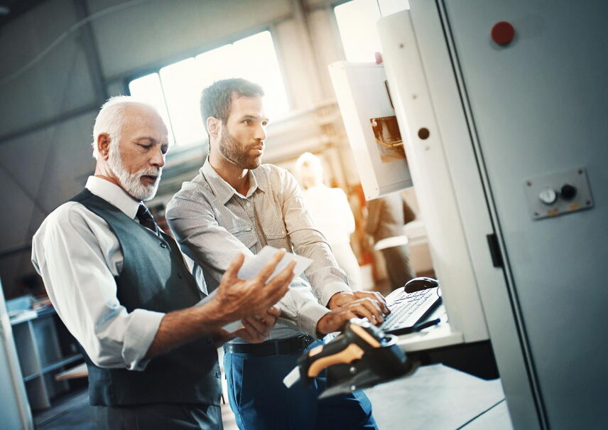 Eine ältere männliche Person mit grauen Haaren und eine jüngere Person stehen zusammen an einem Arbeitsplatz und betrachten ein Blatt und einen Bildschirm.