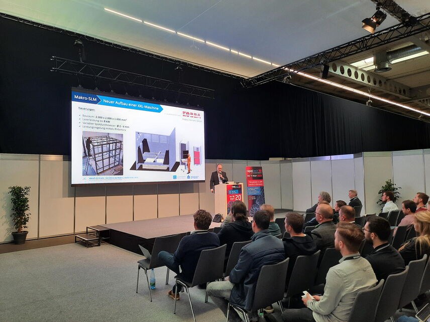 Es ist eine Bühne mit einem Redner zu sehen. Davor sitzen mehrere Personen, die der Präsentation folgen.