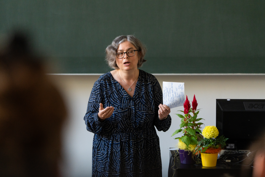 Ein Frau vor einer grünen Tafel, mit einem beschriebenen Blatt Papier in der Hand, die eine Rede hält