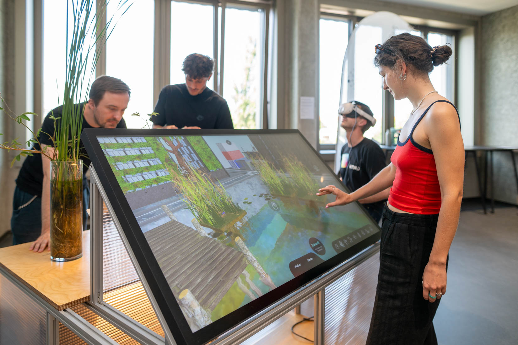 Eine Person bedient den Multitouchscreen, eine weitere die VR-Anwendung und zwei Personen stehen am Tisch des Exponats.