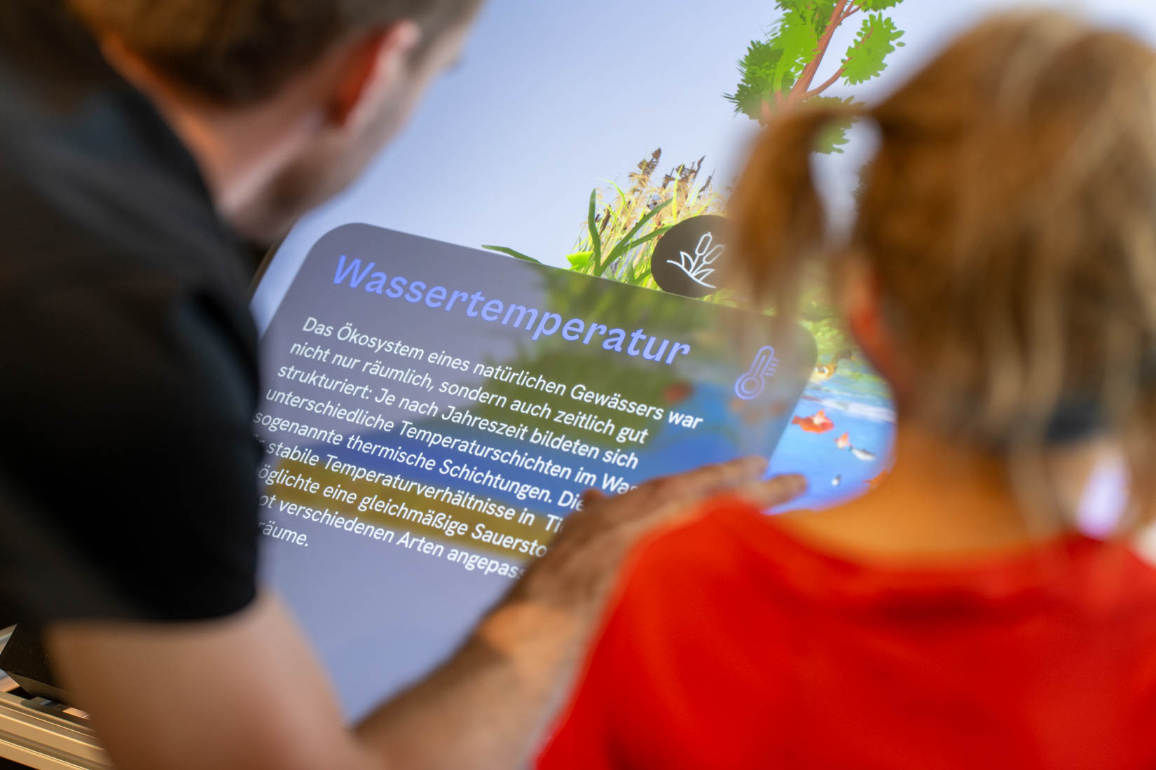 Zwei Personen bedienen den Bildschirm des Exponats, auf dem etwas über die Wassertemperatur eines Gewässers dargestellt wird.