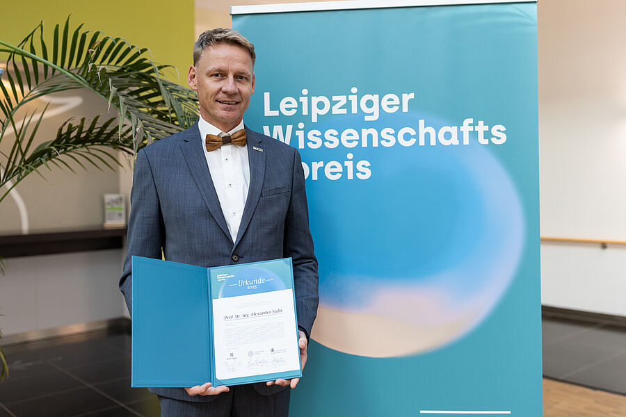 Prof. Dr.-Ing. Alexander Stahr wurde mit dem Leipziger Wissenschatspreis 2025 ausgezeichnet. (Foto: Christian Hüller) Ein Mann hält eine Urkunde und steht vor einem Rollup.