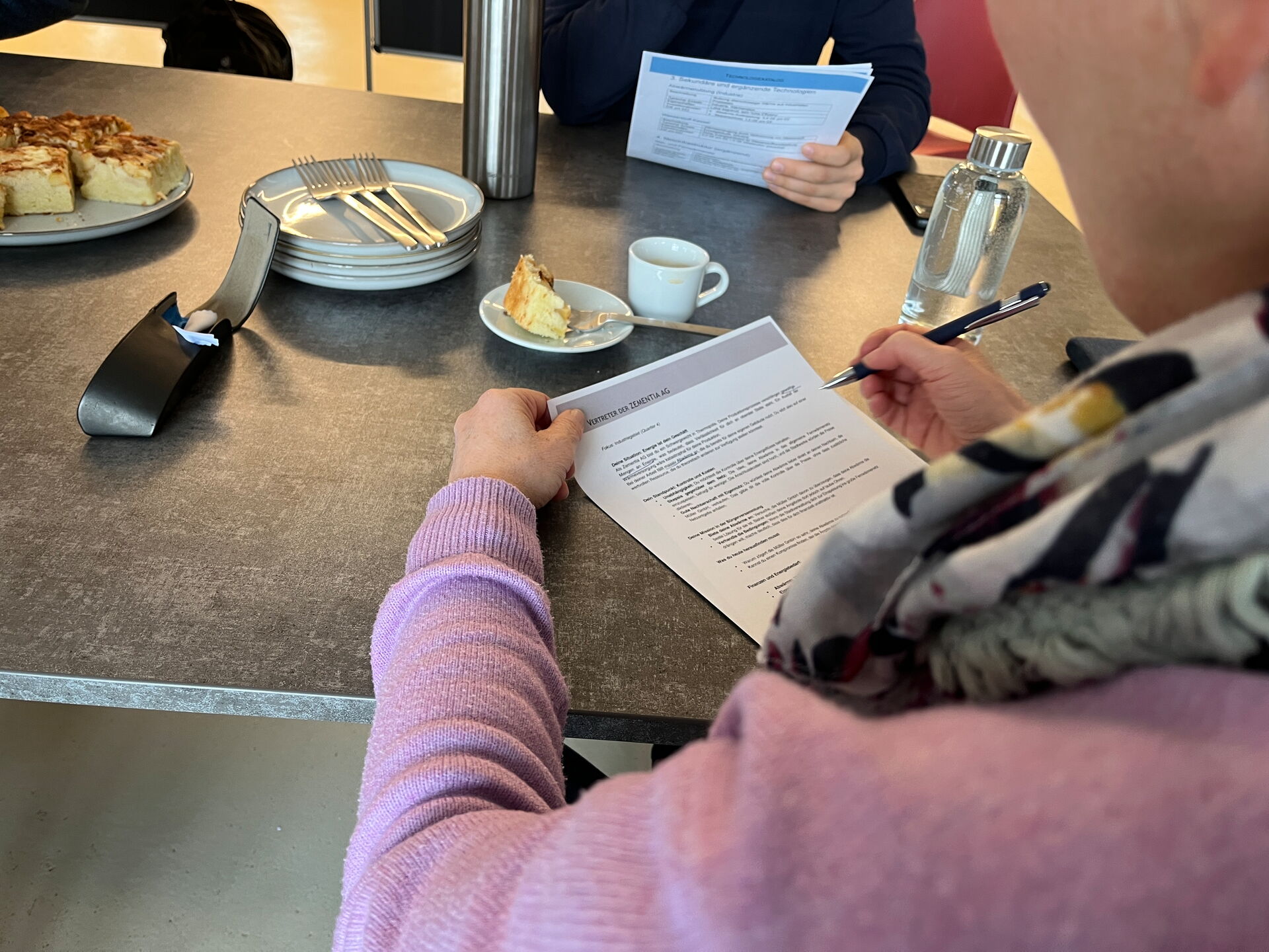 Nahaufnahme: Person macht Notizen auf Rollenblatt; am Tisch stehen Kaffee und Kuchen.