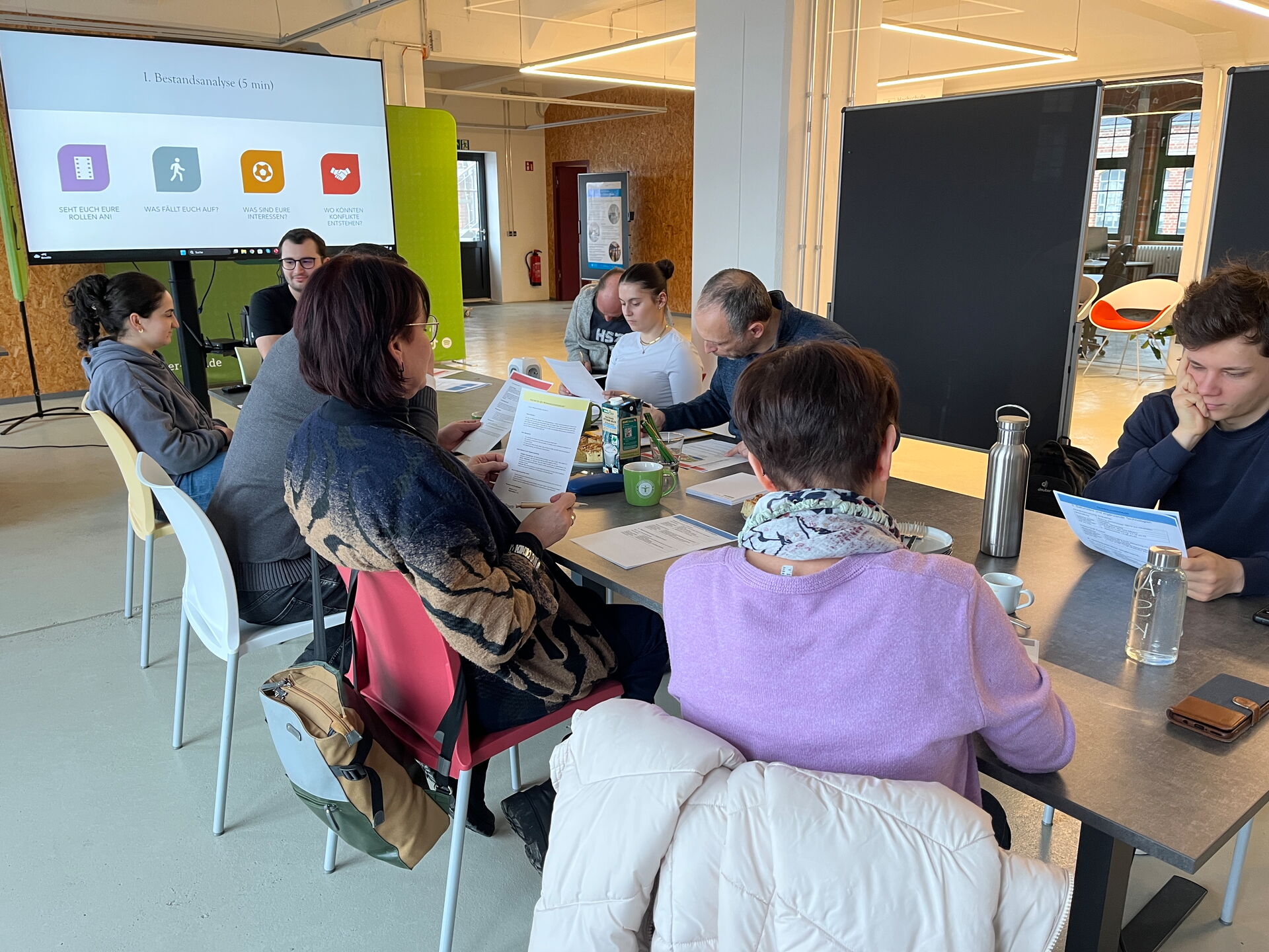 Teilnehmende lesen Rollenblätter am Tisch; Bildschirm zeigt Schritt „Bestandsanalyse“