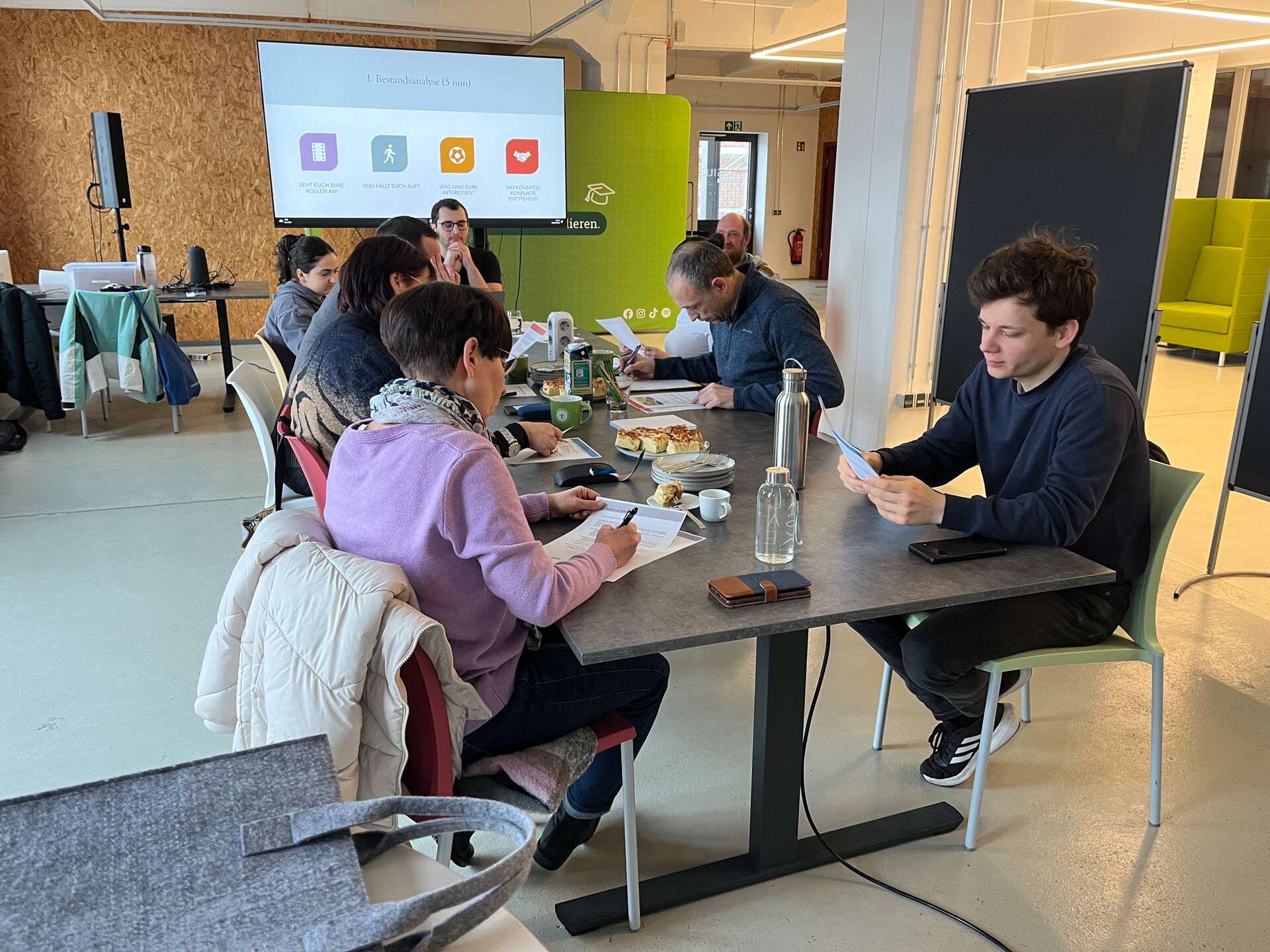 Gruppe arbeitet am Tisch mit Unterlagen; Präsentation zeigt „Bestandsanalyse (5 min)“.