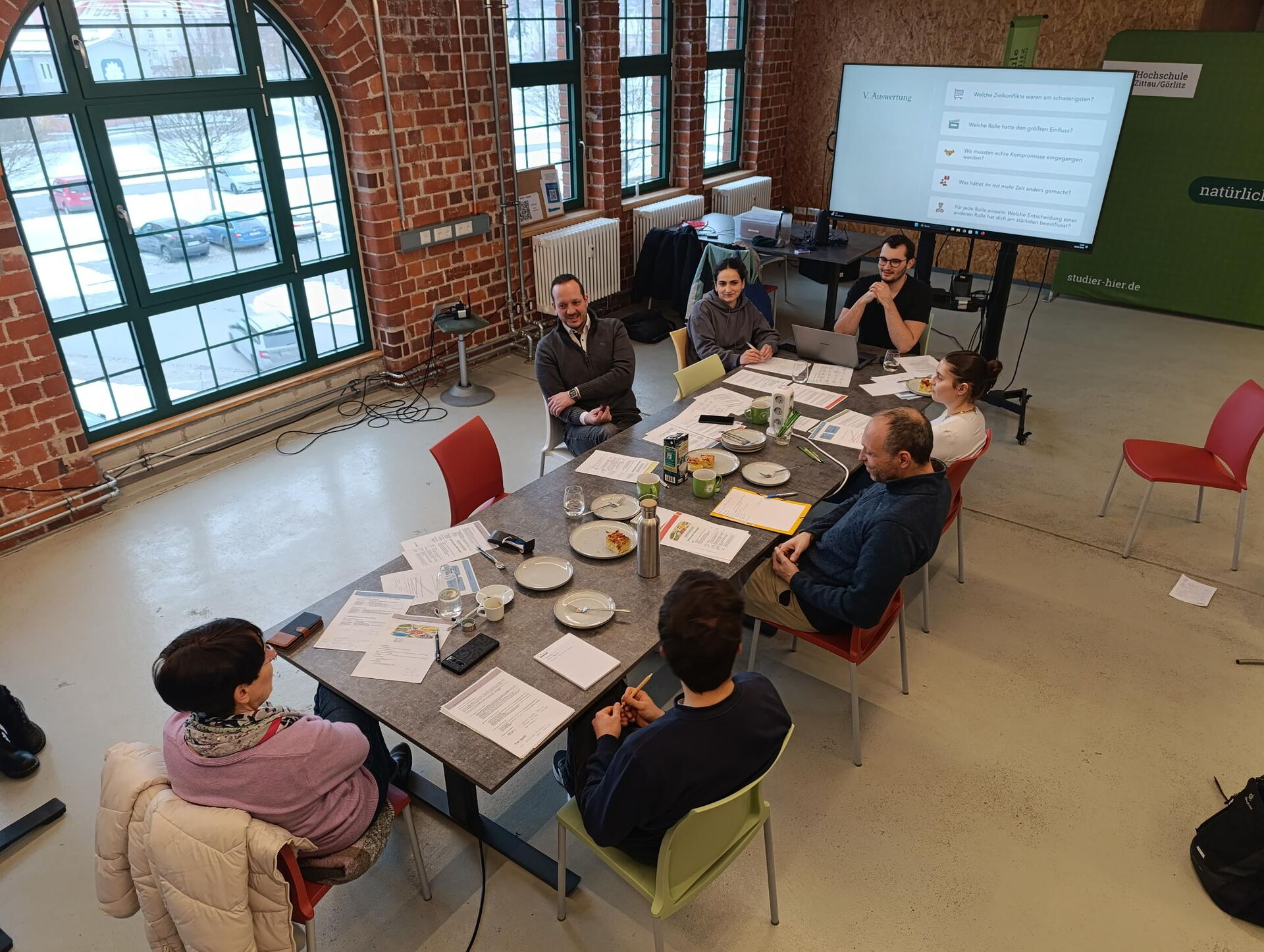 Vogelperspektive auf Tischrunde; auf dem Bildschirm laufen Fragen zur Auswertung.