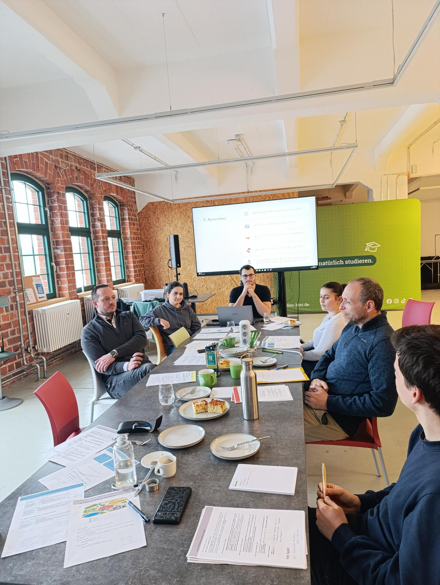 Diskussionsrunde im Planspiel: Teilnehmende am Tisch, Präsentation zur Auswertung im Hintergrund.