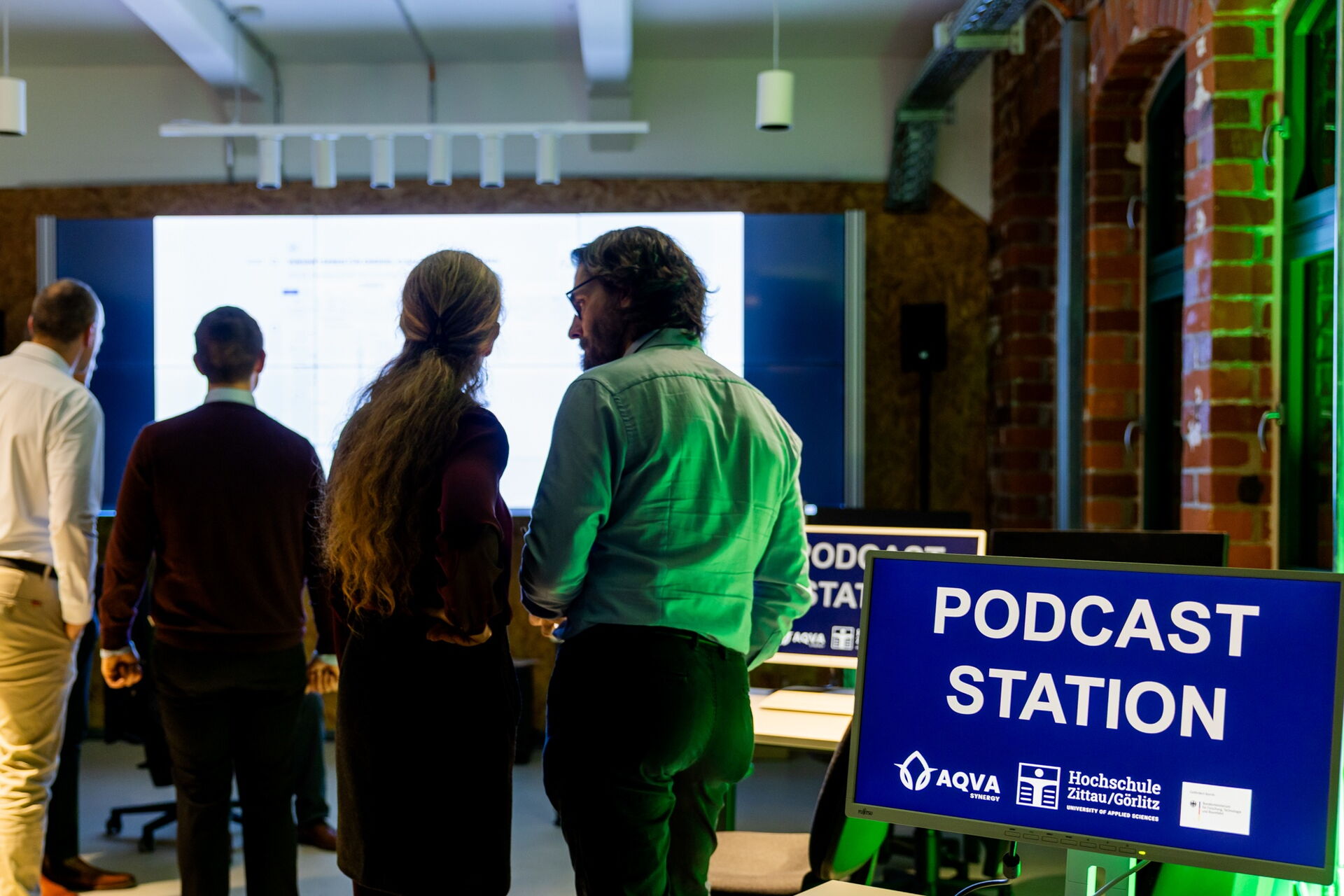Mehrere Personen stehen vor einer großen Leinwand, auf Bildschirmen steht "Podcaststation"