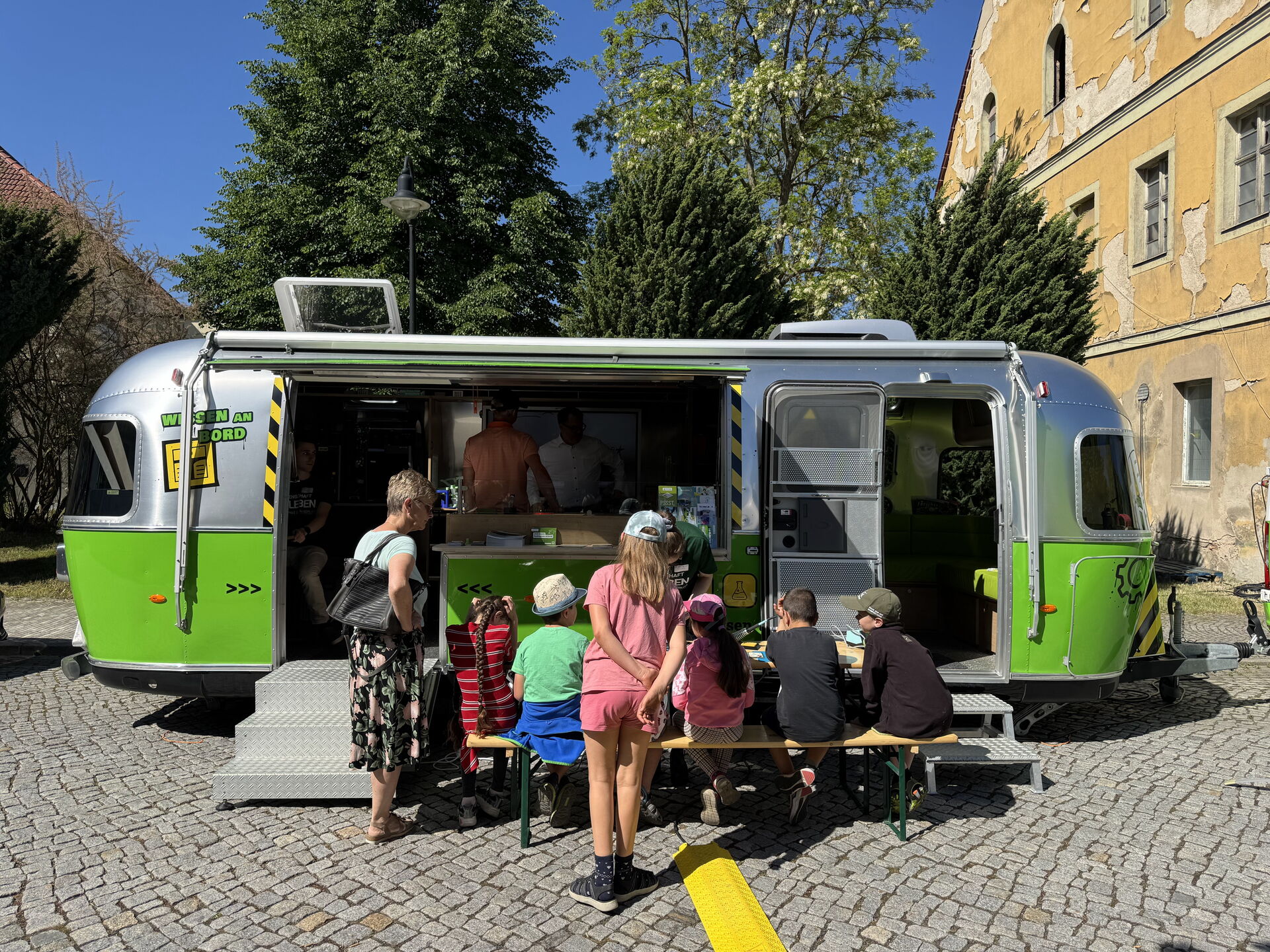 Mehrere Kinder sitzen vor einem grün-silbernen Wissenschaftsanhänger an einem Tisch.