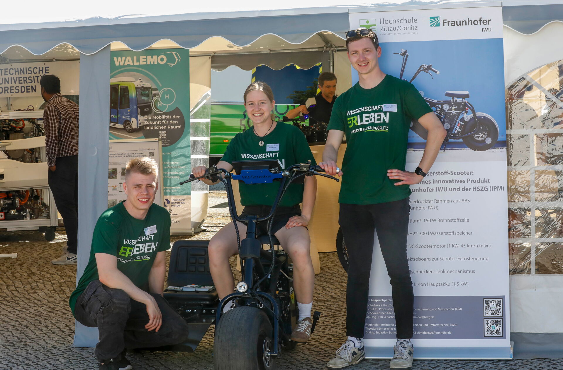 Drei junge Erwachsene präsentieren einen Wasserstoff-Scooter vor Informationsbannern.