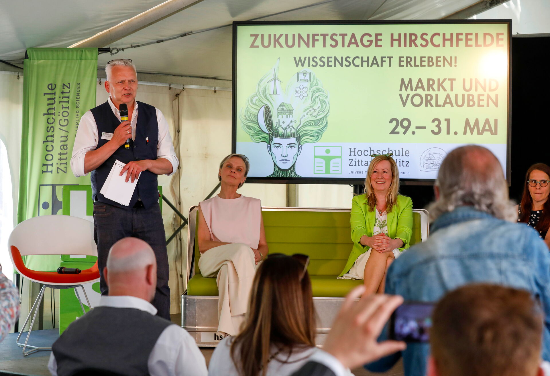 Ein Mann spricht auf der Bühne ins Mikrofon, während drei Frauen hinter ihm sitzen.