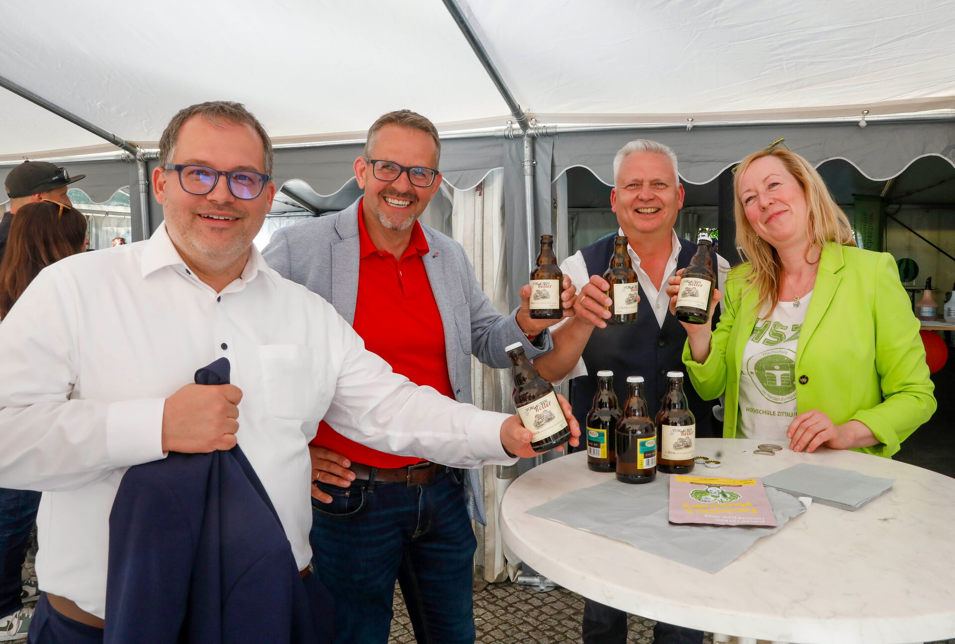 Vier Personen stehen lächelnd an einem Tisch und halten Bierflaschen in die Kamera.