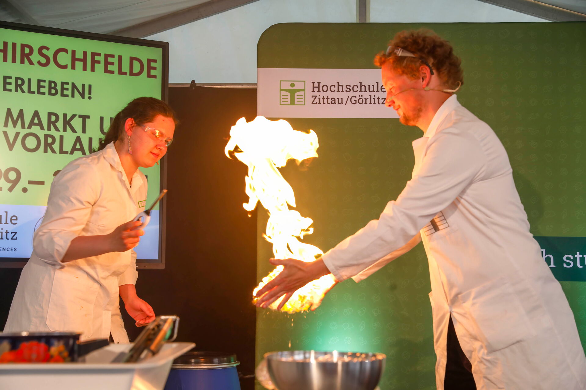 Zwei Personen im Laborkittel führen auf der Bühne ein Experiment mit einer großen Flamme vor.