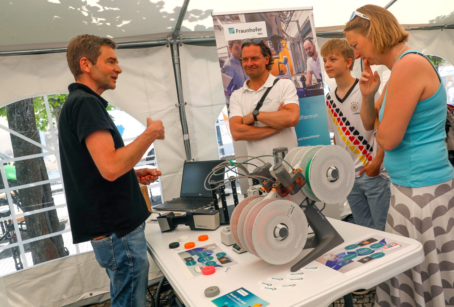 Ein Mann erklärt drei Gästen eine Anlage mit mehreren Materialrollen auf einem Ausstellungstisch.