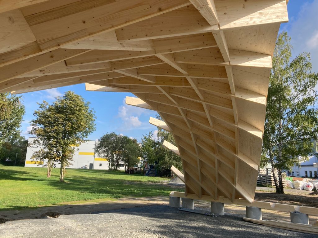Gebogene Holzkonstruktion mit Rippenstruktur, die sich wie ein Dach über den Boden spannt.