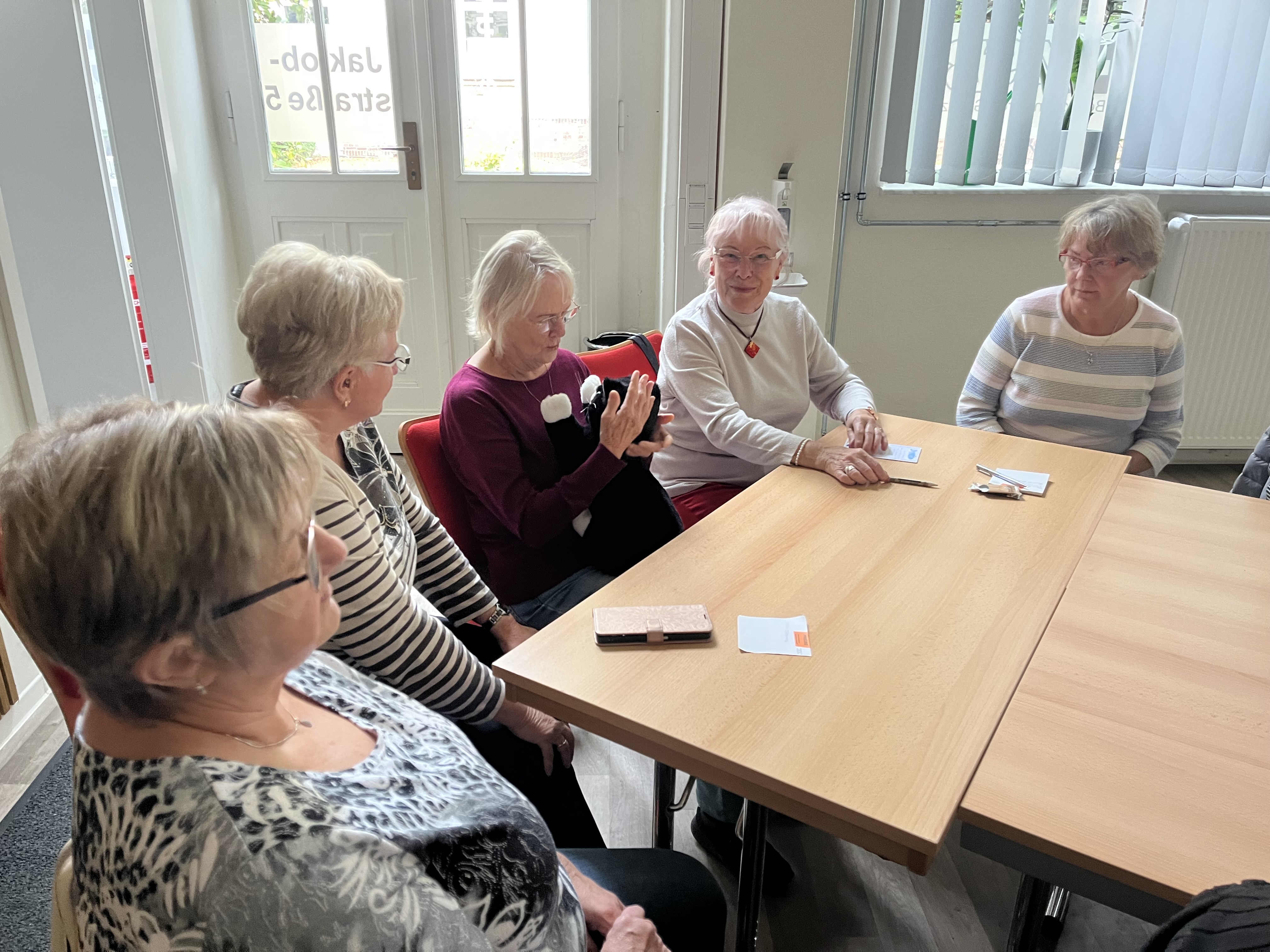 5 Seniorinnen sitzen ein einem Tisch. Eine von Ihnen hat die Roboterkatze JustCat in den Händen.